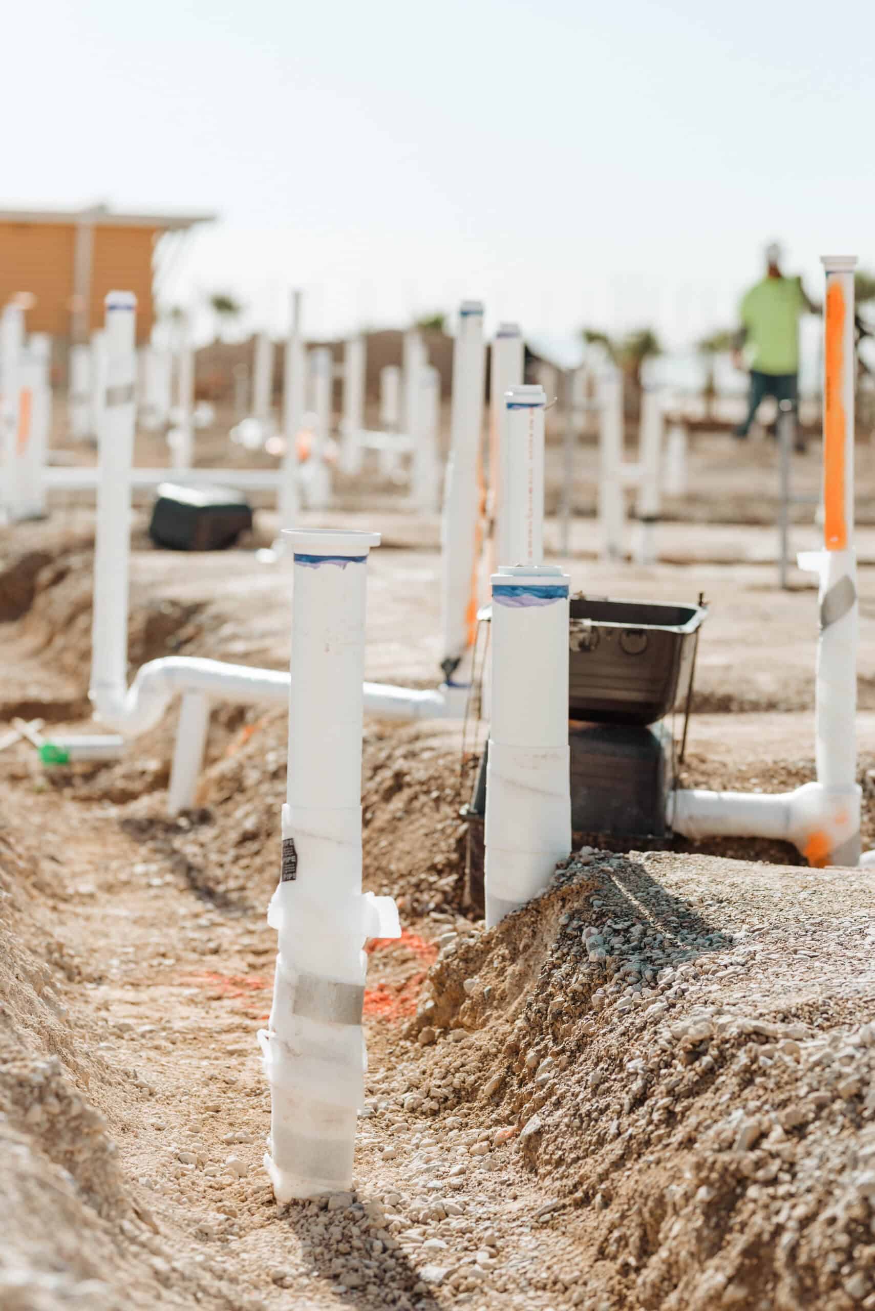 Underground plumbing rough-in with multiple PVC pipe stubs installed across a construction site, prepared for future building connections.