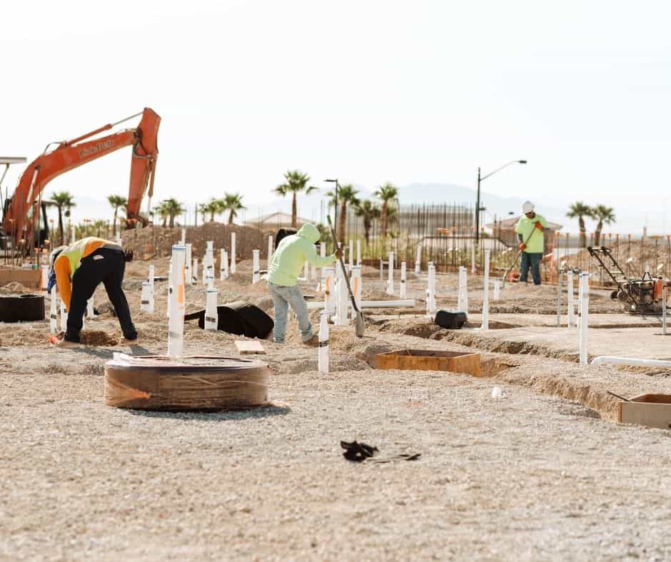 Construction crew installing underground plumbing lines at a residential job site, with multiple pipe stubs visible and an excavator in the background.
