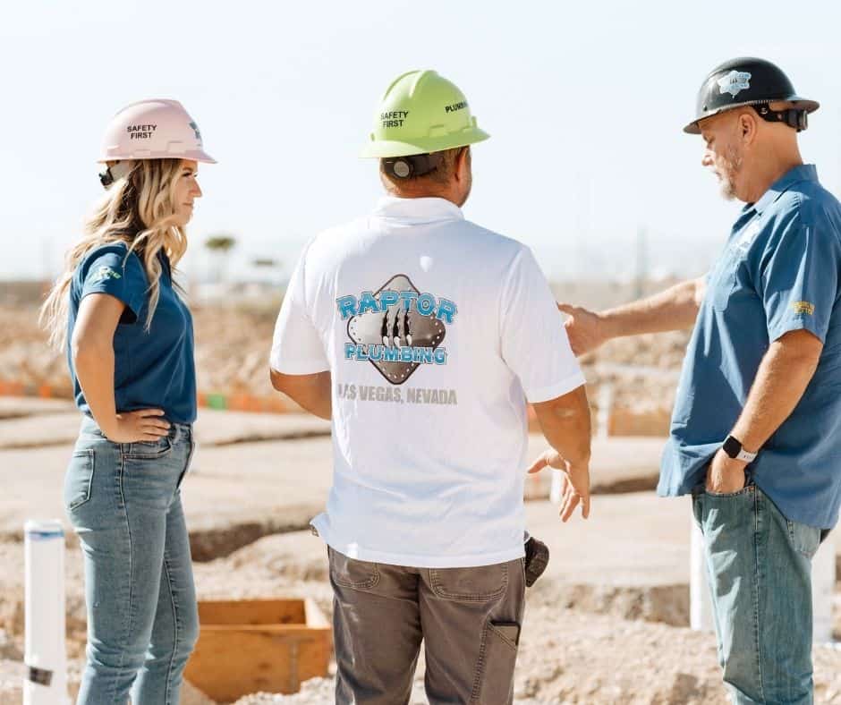 Three construction professionals wearing safety helmets discussing plans on a job site, with visible groundwork and plumbing layout in progress.